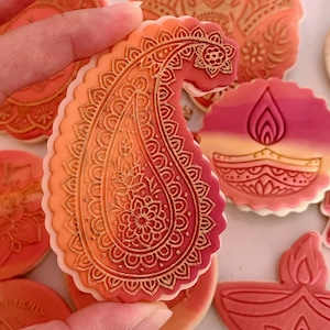 May include: A hand holding a paisley-shaped biscuit with detailed embossed patterns in orange and red hues. Other biscuits with floral and flame designs are visible, all with a similar colour palette and decorative style.
