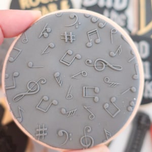 May include: A circular biscuit with a grey surface, decorated with embossed musical notes, treble clefs, and sharp symbols. The biscuit has a light brown edge. Other biscuits with text are visible in the background.