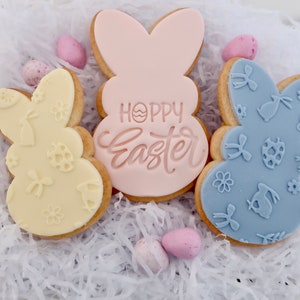 May include: Three Easter bunny-shaped cookies with pastel icing. One is pale yellow with embossed designs, another is light pink with "Hoppy Easter" text, and the third is light blue with embossed patterns. Small pastel eggs are scattered around.