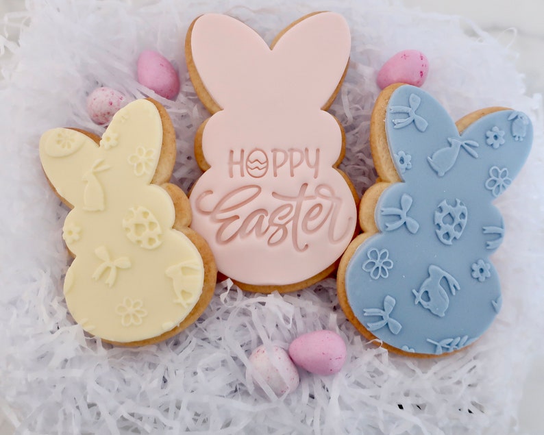 May include: Three Easter bunny-shaped cookies with pastel icing. One is pale yellow with embossed designs, another is light pink with "Hoppy Easter" text, and the third is light blue with embossed patterns. Small pastel eggs are scattered around.