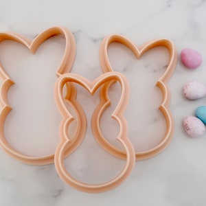 May include: Three light peach-colored bunny-shaped cookie cutters in varying sizes. The cutters are made of plastic and have a smooth, rounded edge. Several pastel-colored Easter egg candies are scattered nearby, suggesting a springtime theme.