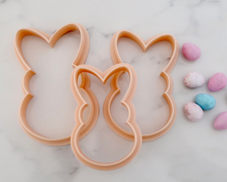 May include: Three light peach-colored bunny-shaped cookie cutters in varying sizes. The cutters are made of plastic and have a smooth, rounded edge. Several pastel-colored Easter egg candies are scattered nearby, suggesting a springtime theme.