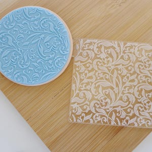 May include: A light blue, circular cookie stamp with a floral design next to a clear, square stamp with the same pattern. Both stamps are on a wooden surface. The floral design features swirling leaves and vines.