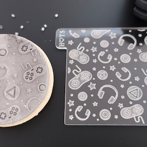 May include: A round cookie with a grey embossed pattern of gaming icons, including controllers, headphones, stars, and geometric shapes. Beside it is a clear acrylic stamp with the same design. The stamp has the text "HOTS".