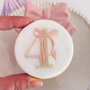 Könnte beinhalten: Ein runder Keks mit weißem Zuckerguss, verziert mit einer hellrosa Schleife und der Zahl 4 in hellem Beige. Eine weitere rosa Schleife ist auf einem weißen Teller im Hintergrund zu sehen. Der Keks wird von einer Hand gehalten.