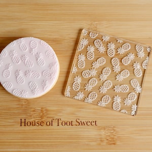 May include: A round, light pink cookie with a white pineapple pattern next to a clear, square acrylic stamp with the same pineapple design. The text "House of Toot Sweet" is visible.