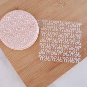 May include: A light pink, circular cookie with a white bow pattern, alongside a clear, square stencil also featuring a bow pattern. Both are on a wooden board. The bows are uniformly arranged.