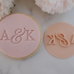 May include: A light pink cookie with the letters "A & K" imprinted on it, next to a light pink circular stamp with the letters "K & A". The cookie has a light brown edge. Soft pink flowers are in the background.