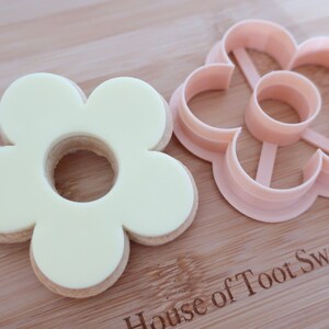 May include: A flower-shaped cookie cutter and a decorated biscuit sit on a wooden surface. The biscuit is light yellow with a flower shape and a hole in the centre. The cutter is light pink and has the same flower shape.
