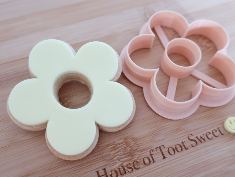 May include: A flower-shaped cookie cutter and a decorated biscuit sit on a wooden surface. The biscuit is light yellow with a flower shape and a hole in the centre. The cutter is light pink and has the same flower shape.