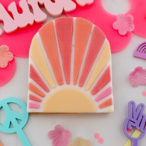 May include: A decorative, retro-style sunburst design in shades of orange, peach, and yellow. The design is on a white background, with a semi-circular shape. Surrounding the sunburst are pink flowers, peace signs, and a rainbow.