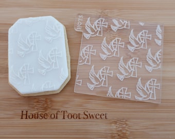 May include: A white frosted cookie with an embossed dove and cross design, next to a clear acrylic stamp with the same pattern. The cookie is on a wooden surface, with the text "House of Toot Sweet" engraved below.