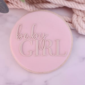 May include: A round cookie with a light pink frosting. The words "baby GIRL" are written in a light gray, elegant font across the center of the cookie. The cookie is a dessert item.
