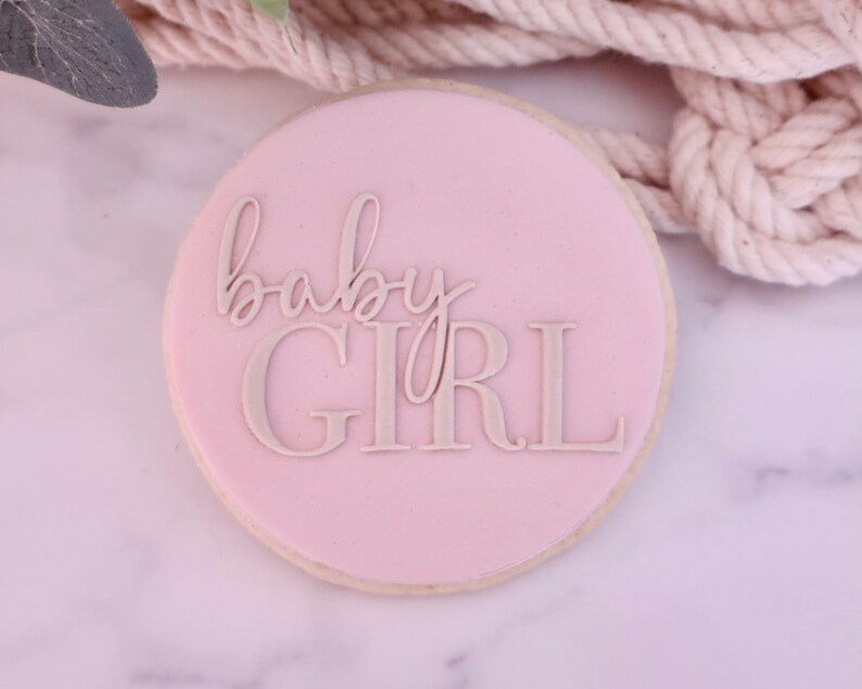 May include: A round cookie with a light pink frosting. The words "baby GIRL" are written in a light gray, elegant font across the center of the cookie. The cookie is a dessert item.