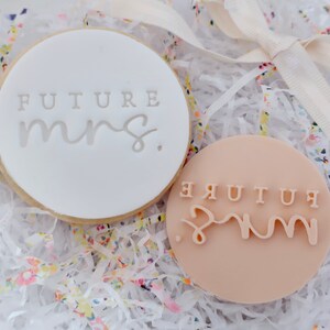 Könnte beinhalten: Zwei runde Kekse, einer weiß und einer hellpfirsichfarben, mit dem Text "FUTURE mrs." eingeprägt. Die Kekse werden in einer Geschenkbox mit buntem Papier und einem cremefarbenen Band präsentiert.