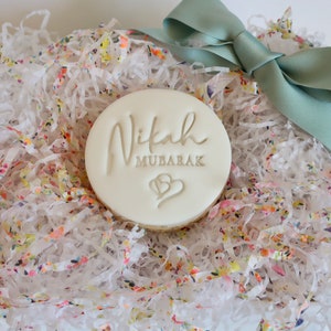 Puede incluir: Una galleta redonda y blanca con las palabras "Nikah Mubarak" y dos corazones, sobre un lecho de papel triturado colorido. Una cinta verde claro atada en un lazo a&ntilde;ade un toque decorativo a la presentaci&oacute;n del regalo.