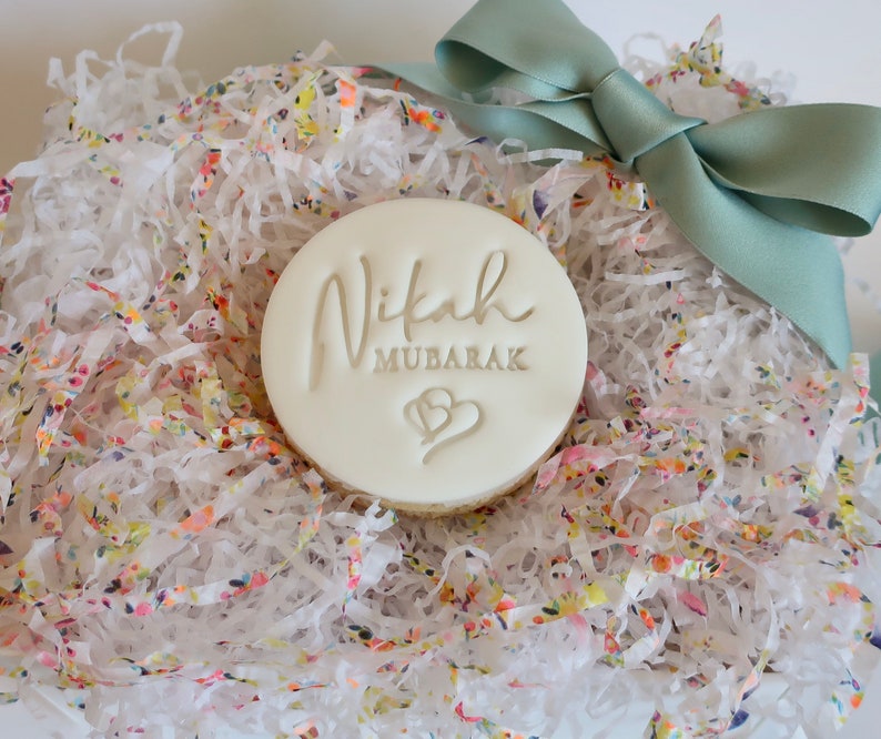 Puede incluir: Una galleta redonda y blanca con las palabras "Nikah Mubarak" y dos corazones, sobre un lecho de papel triturado colorido. Una cinta verde claro atada en un lazo a&ntilde;ade un toque decorativo a la presentaci&oacute;n del regalo.