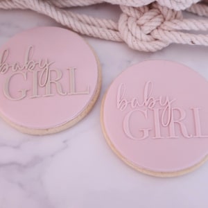May include: Two round cookies with a light pink frosting. The cookies have the words "baby GIRL" in a lighter shade of pink. The cookies are on a white marble surface.