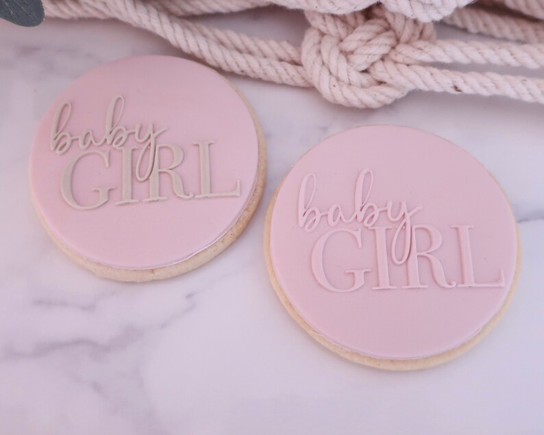 May include: Two round cookies with a light pink frosting. The cookies have the words "baby GIRL" in a lighter shade of pink. The cookies are on a white marble surface.
