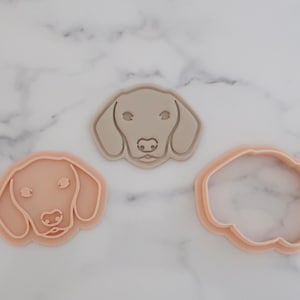 May include: Three dog-shaped cookie cutters in shades of beige. Two cutters depict a dog's head, one with facial details and the other an outline. The third cutter is the outline of the dog's head.