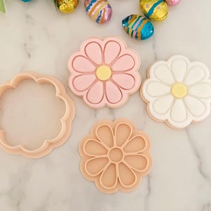 May include: A set of flower-shaped cookie cutters and stamps in light peach plastic. The set includes a plain circular cutter with scalloped edges, a stamp with a pink flower design, a stamp with a white flower design, and a peach-coloured stamp.
