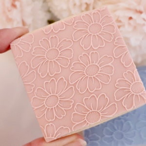 May include: A square, light pink cookie stamp with a repeating daisy pattern. The daisy design is raised and in a lighter pink color. A second, blue stamp with a similar design is visible in the background.