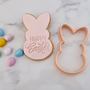 May include: A light pink Easter bunny-shaped cookie with the words "Hoppy Easter" in a cursive font. A matching bunny-shaped cookie cutter is next to the cookie. Colorful speckled Easter eggs are scattered around the cookie.