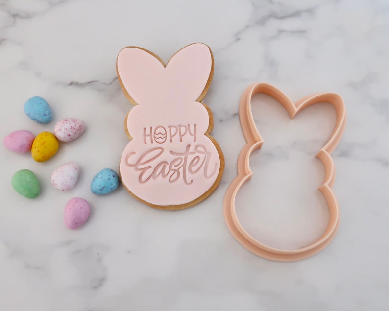 May include: A light pink Easter bunny-shaped cookie with the words "Hoppy Easter" in a cursive font. A matching bunny-shaped cookie cutter is next to the cookie. Colorful speckled Easter eggs are scattered around the cookie.