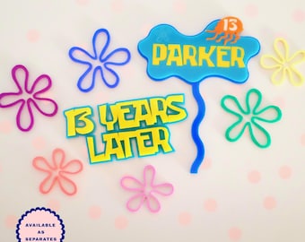 May include: A blue cake topper with the text PARKER and a small orange jellyfish icon, a yellow cake topper with the text 13 YEARS LATER, and several colorful flower-shaped outlines in purple, blue, yellow, green, pink, and coral colors.