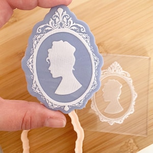 May include: A blue and white cameo-style cookie cutter featuring a woman's profile within an oval frame. A hand holds the cutter, with a clear plastic template and a pink cutter base also visible.