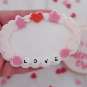 May include: A white, heart-shaped cookie decorated with a pink and white beaded bracelet design. The bracelet has pink stars, a red heart, and the word "LOVE" spelled out in black letters. A second cookie is visible in the background.