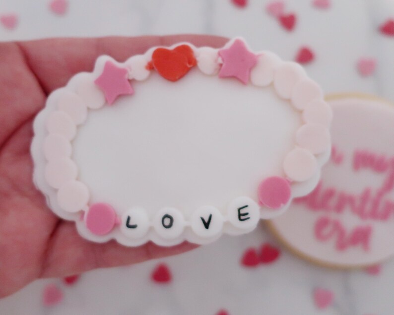 May include: A white, heart-shaped cookie decorated with a pink and white beaded bracelet design. The bracelet has pink stars, a red heart, and the word "LOVE" spelled out in black letters. A second cookie is visible in the background.