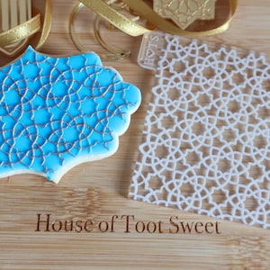May include: A light blue cookie with a gold geometric design, beside a clear stencil with a matching pattern. Gold ribbon and a gold decoration are in the background. The text "House of Toot Sweet" is at the bottom.