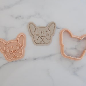May include: Three French Bulldog face-shaped cookie cutters in shades of beige and tan. The cutters are made of plastic and are designed to cut out the shape of a French Bulldog's face. The cutters are arranged on a white marble surface.
