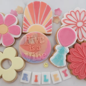 May include: A selection of decorated sugar biscuits with a retro theme. The biscuits feature flower shapes, a sunburst design, a balloon, and the phrase "Five is a Vibe." The name "Riley" is spelled out in individual letter biscuits. Colours include pink, orange, yellow, and blue.