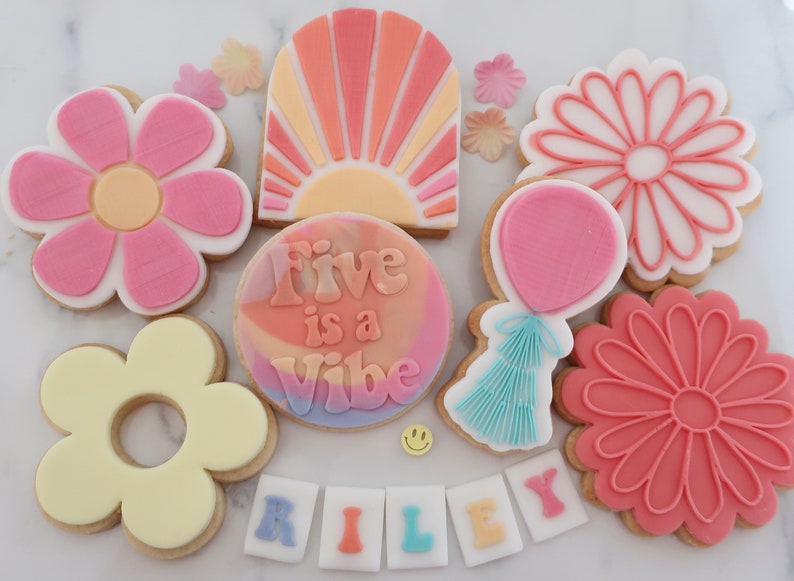 May include: A selection of decorated sugar biscuits with a retro theme. The biscuits feature flower shapes, a sunburst design, a balloon, and the phrase "Five is a Vibe." The name "Riley" is spelled out in individual letter biscuits. Colours include pink, orange, yellow, and blue.