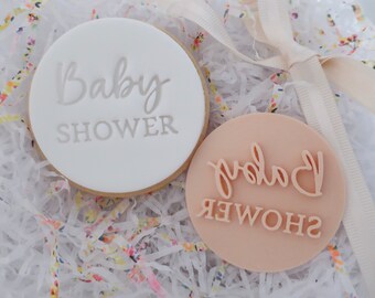 May include: A round white cookie with the embossed text Baby Shower in a light gray color, positioned next to a round peach-colored plastic stamp featuring the raised text Baby Shower in a script and block font combination.