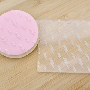 Könnte beinhalten: Ein rosa, runder Keks mit einem Blitzmuster, neben einem transparenten, quadratischen Stempel mit dem gleichen Design. Der Keks liegt auf einer hellen, runden Basis. Der Stempel besteht aus transparentem Kunststoff.