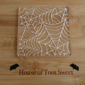 May include: A clear, square coaster with a detailed spiderweb design. The coaster is placed on a wooden surface with the text "House of Toot Sweet" and two black bat cutouts. Ideal for Halloween.