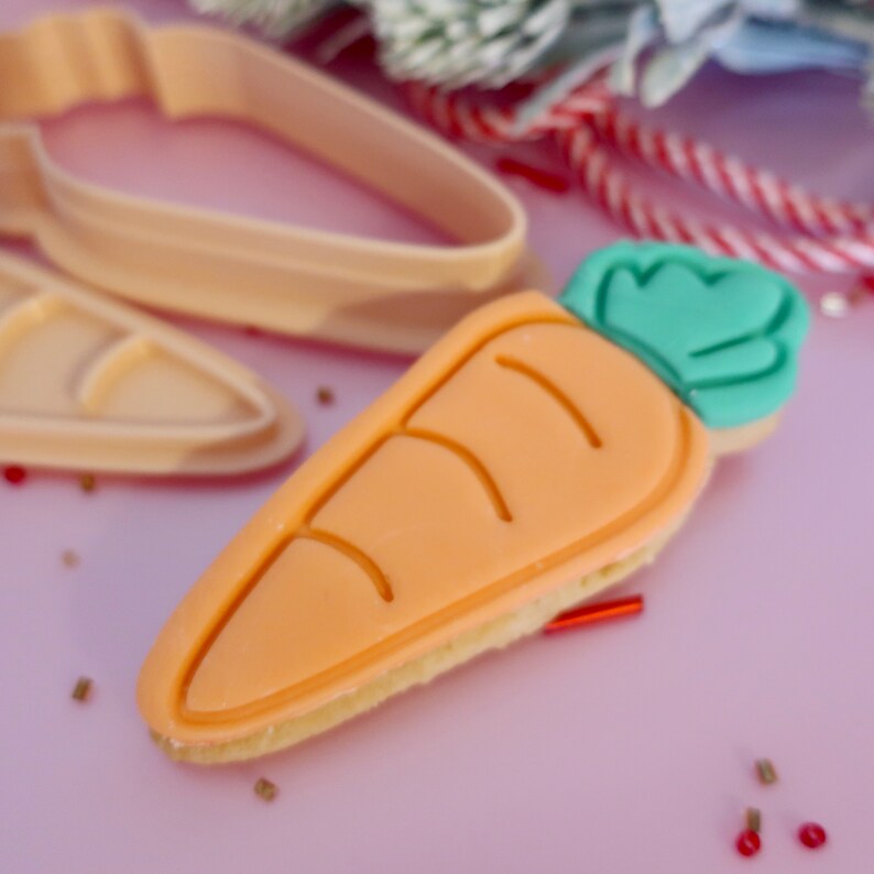 May include: A carrot-shaped biscuit with orange icing and green icing for the leaves. The biscuit is on a light pink surface, with a carrot-shaped biscuit cutter in the background. The biscuit has a light brown base.