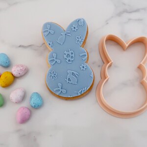 May include: A light blue Easter bunny-shaped cookie with embossed designs, next to a matching cookie cutter. Colorful speckled candy eggs in pink, yellow, blue, and green are scattered nearby. The cookie features bunny, egg, and floral patterns.