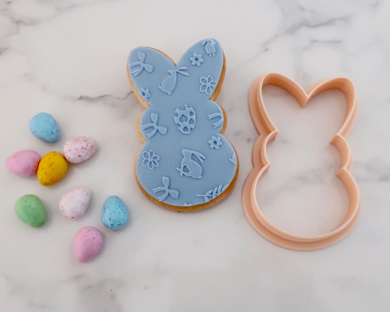 May include: A light blue Easter bunny-shaped cookie with embossed designs, next to a matching cookie cutter. Colorful speckled candy eggs in pink, yellow, blue, and green are scattered nearby. The cookie features bunny, egg, and floral patterns.