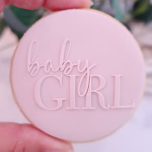 May include: A round cookie with a light pink frosting, featuring the words "baby GIRL" in a raised, elegant font. The cookie has a light brown edge, and the overall design suggests a baby shower or gender reveal theme.