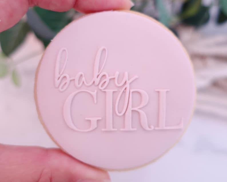 May include: A round cookie with a light pink frosting, featuring the words "baby GIRL" in a raised, elegant font. The cookie has a light brown edge, and the overall design suggests a baby shower or gender reveal theme.