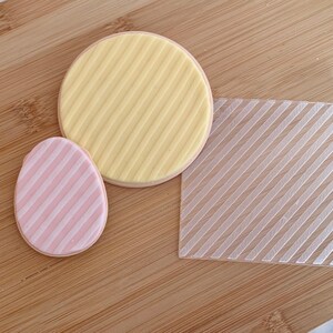 May include: An Easter-themed image featuring two decorated cookies and a clear plastic stencil. One cookie is egg-shaped with pink and white stripes, and the other is round with yellow and white stripes. The stencil has diagonal lines.