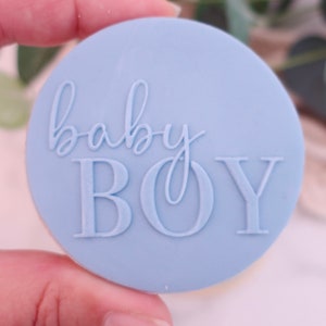May include: A light blue, circular stamp with the words "baby BOY" embossed in a script and block font. The stamp is held in a hand, suggesting its use for crafting or decorating baby-themed items.
