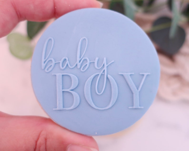 May include: A light blue, circular stamp with the words "baby BOY" embossed in a script and block font. The stamp is held in a hand, suggesting its use for crafting or decorating baby-themed items.