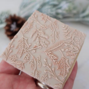 May include: A square, light beige biscuit featuring an embossed, intricate pattern of holly leaves, pine needles, and small berries across the entire surface. The design is raised and detailed, covering the flat top of the baked treat.