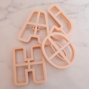Könnte beinhalten: Vier hellpfirsichfarbene Ausstecher in Form der Buchstaben A, H, J und O. Die Ausstecher sind aus Kunststoff und auf einer hellen Marmoroberfläche angeordnet. Die Buchstaben sind in einer serifenlosen Schriftart.