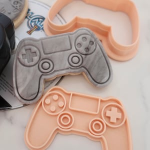 May include: A gray cookie shaped like a video game controller with a marble effect, next to two peach-colored plastic cookie cutters in the same shape. The cookie has a plus sign, circles, and buttons. The cookie cutters are on a white surface.